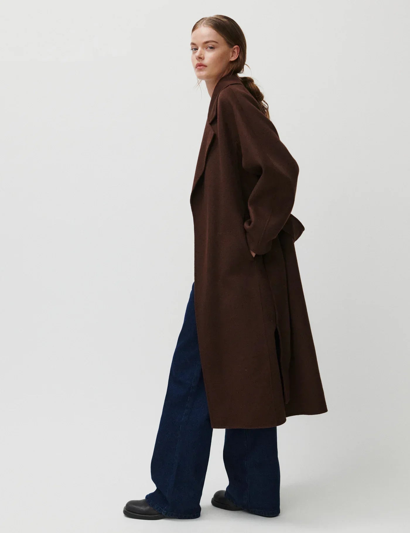 Mørkebrun frakke med bælte, reverskrave, knælængde og slids bagpå. Båret af en person i sideprofil.