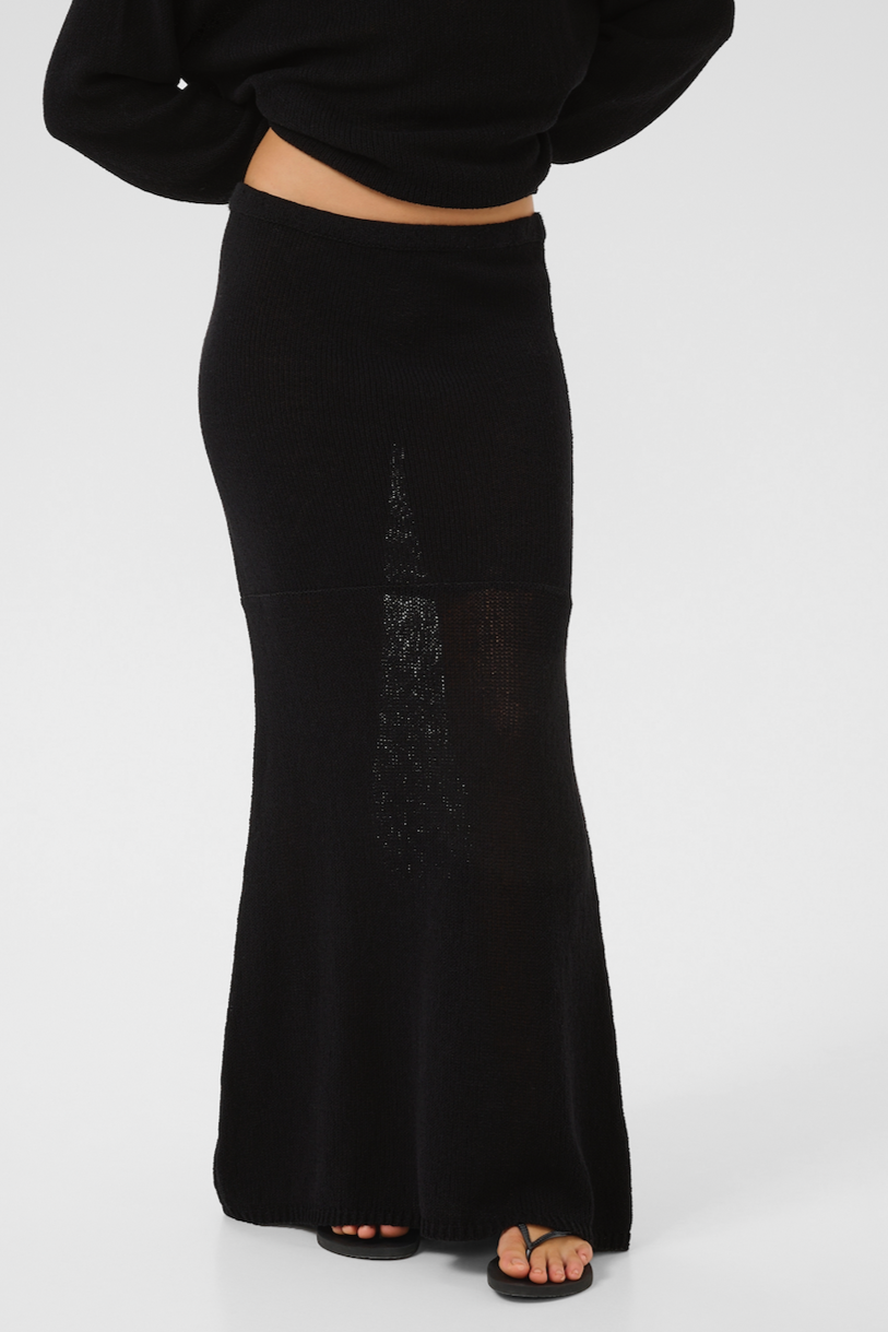 Lang, sort strikket nederdel, tætsiddende og når til fødderne, båret med sorte sandaler.