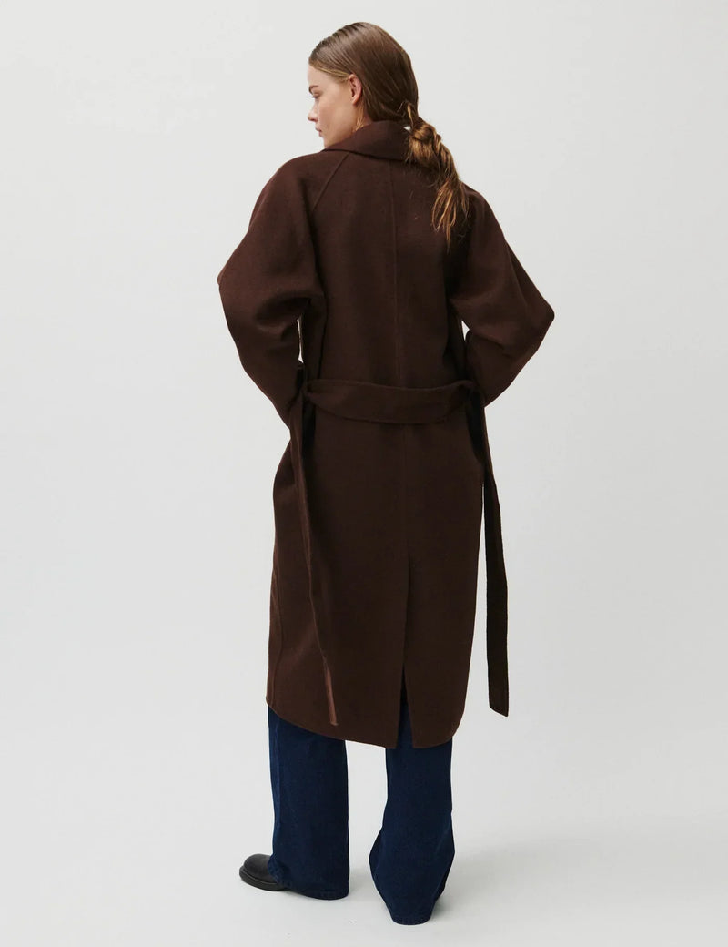 Kvinde iført mørkebrun frakke med bælte og slids bagpå, set bagfra. Frakken er knælængde.