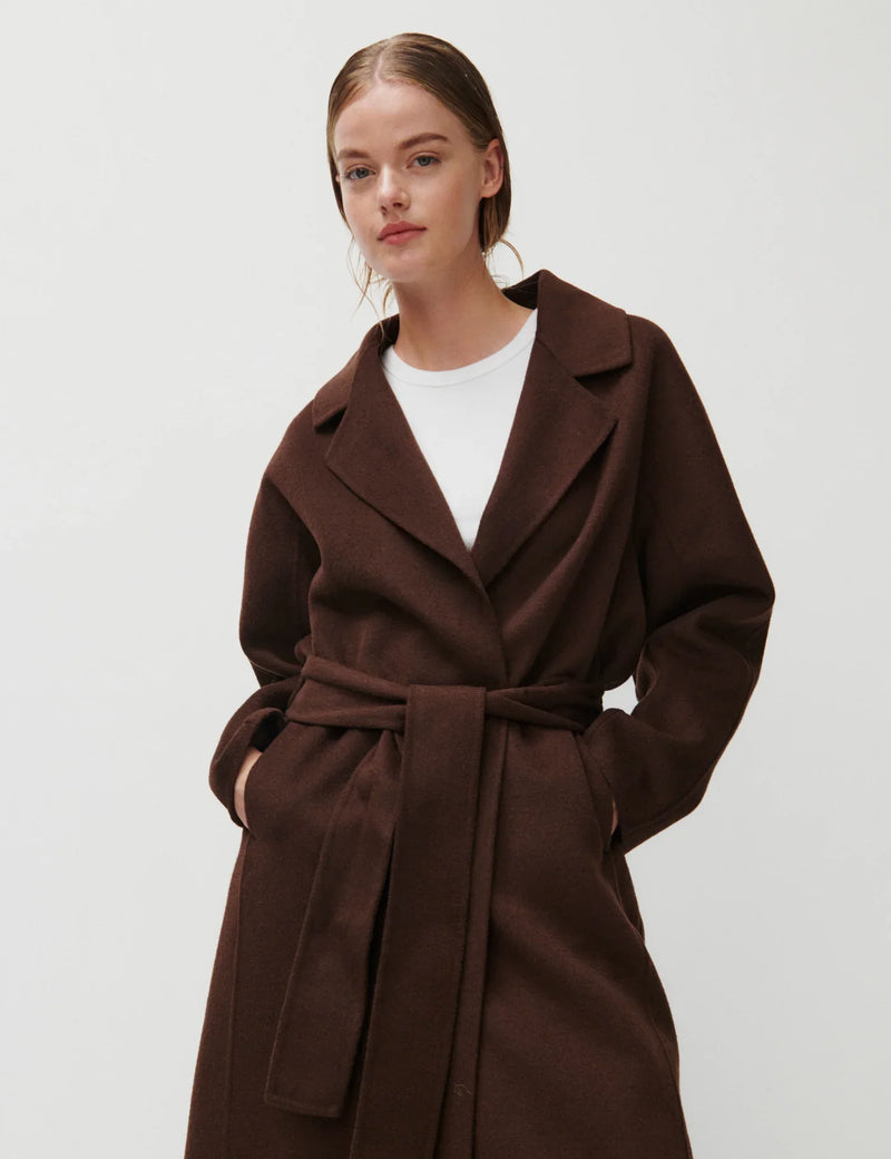 Kvinde i mørkebrun frakke med bælte og klassisk reverskrave. Frakken er knælængde og elegant designet.