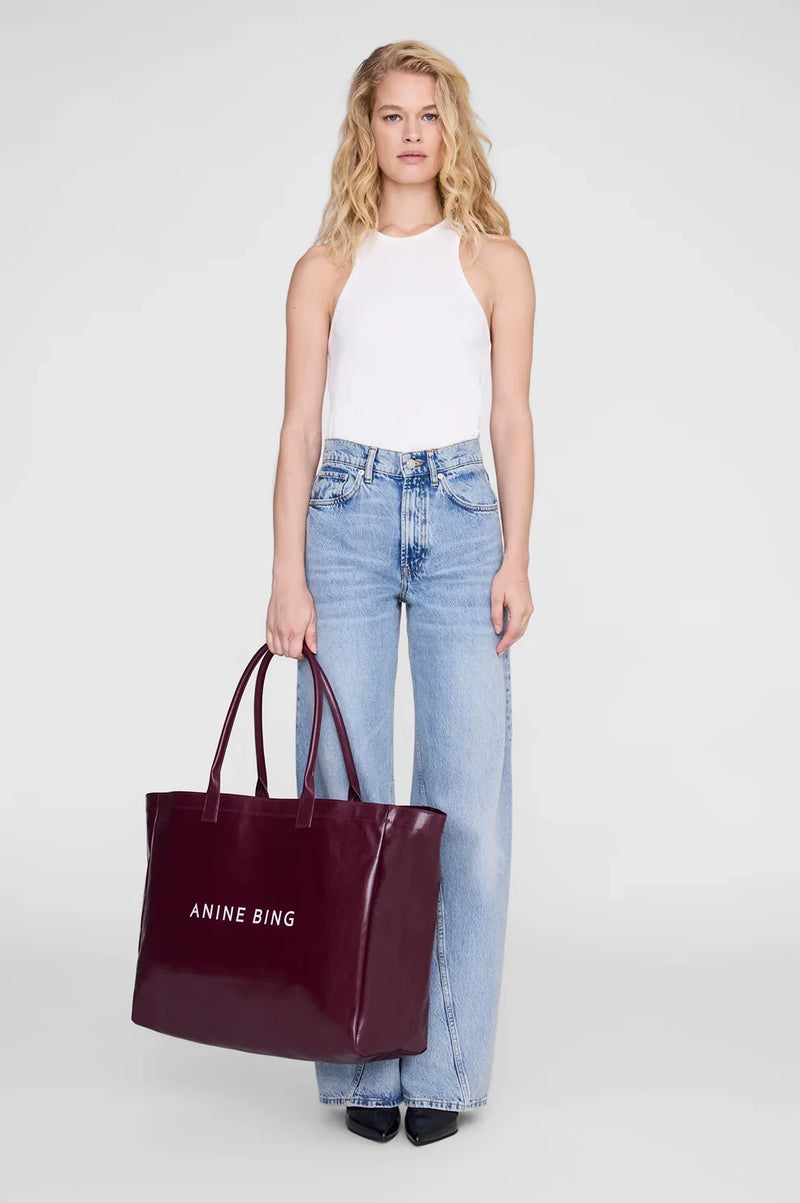 ANINE BING Leo Coated Canvas Tote i dyb bordeaux (Fig) med stort kontrastfarvet logo på fronten og lange skulderstropper hvor en kvinde holder tasken