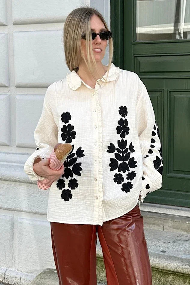 Skjorte med sorte blomster, bølget krave, knapper foran. Person med brød foran grøn dør.