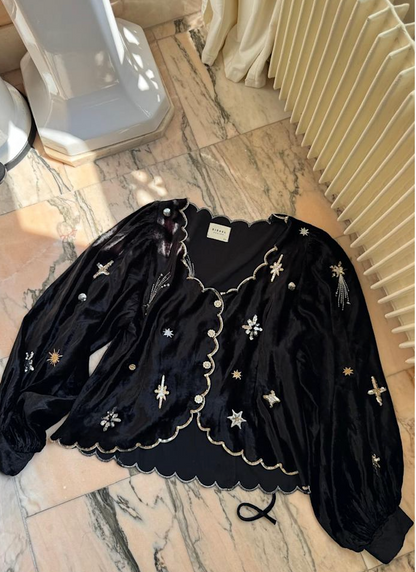 Sort bluse med skinnende stof, broderede stjerner, bølget kant og knapper, liggende på marmor.