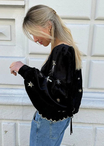 Model i sort bluse med broderede stjerner, bølget kant, knapper foran, og blå jeans.