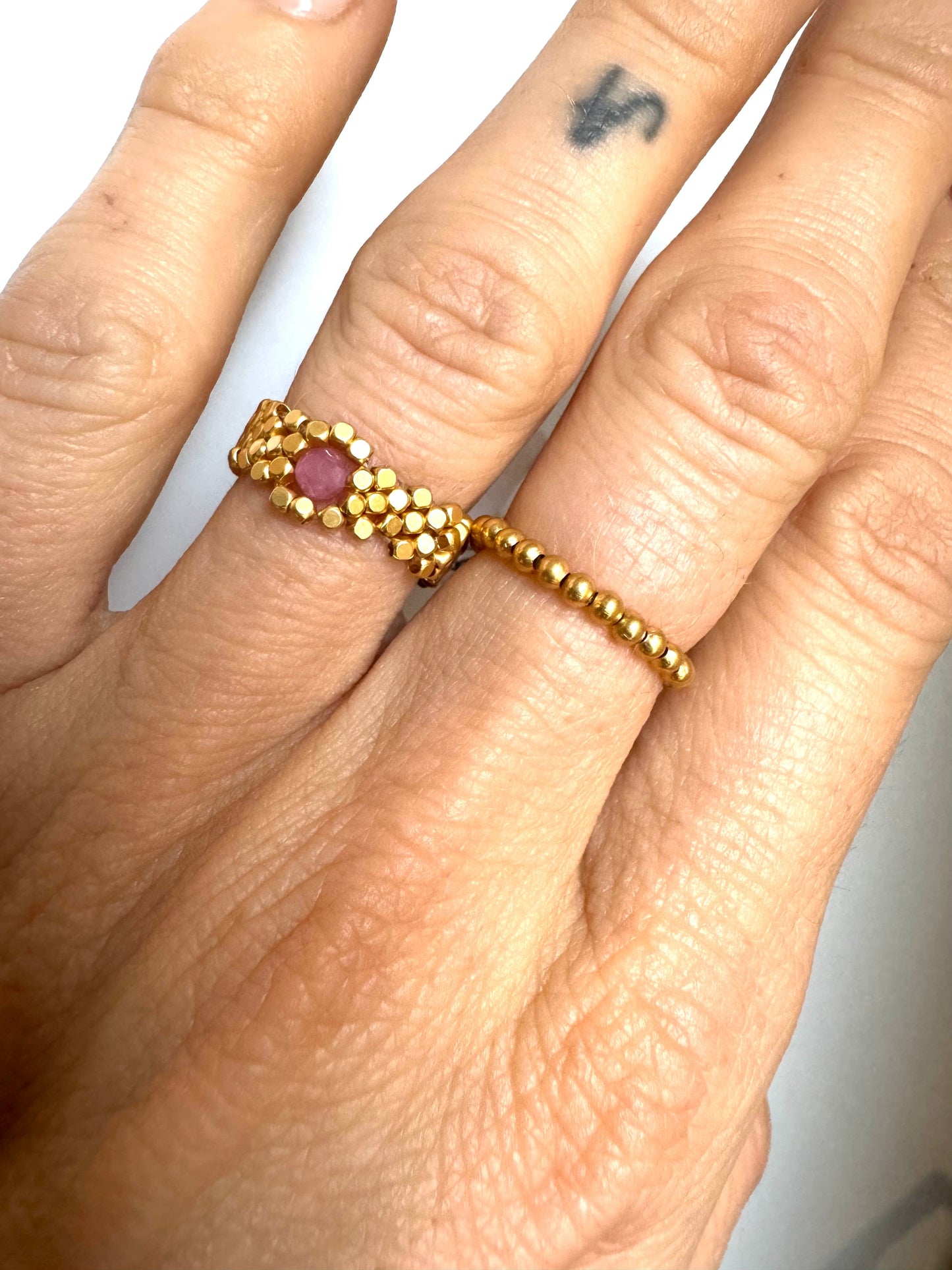 Hånd med guldbelagt ring af små kugler og en rød sten i midten, enkel og tynd i designet.