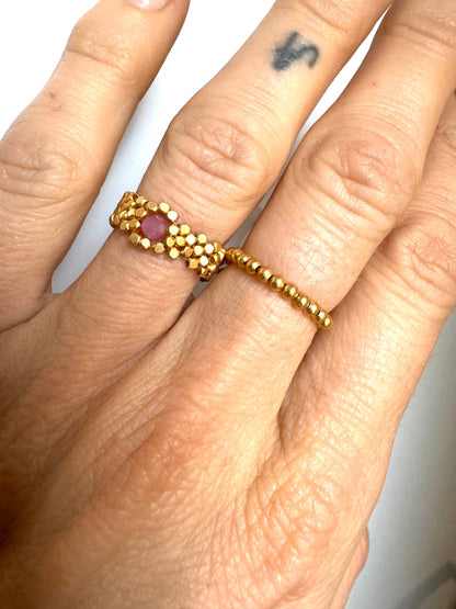 Hånd med guldbelagt ring af små kugler og en rød sten i midten, enkel og tynd i designet.