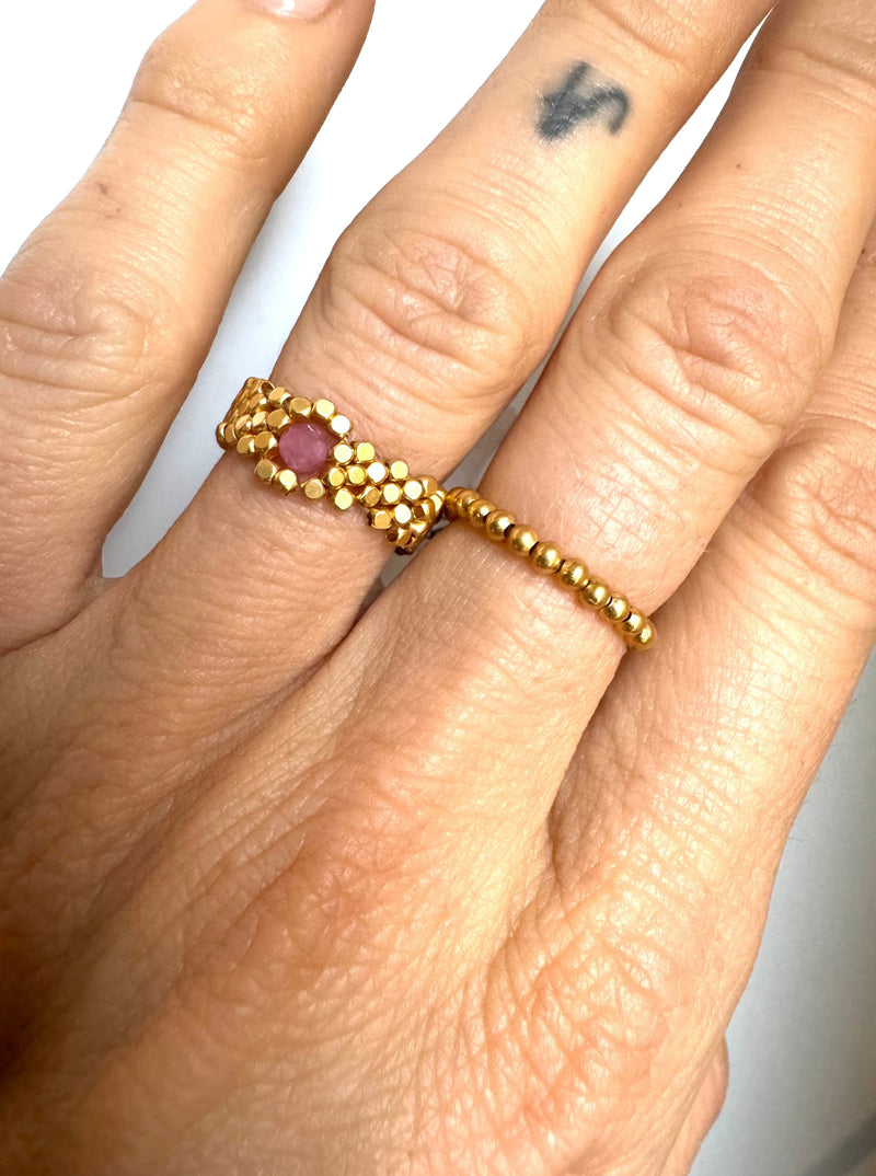 Hånd med guldbelagt ring, lyserød sten og små gyldne perler på finger.